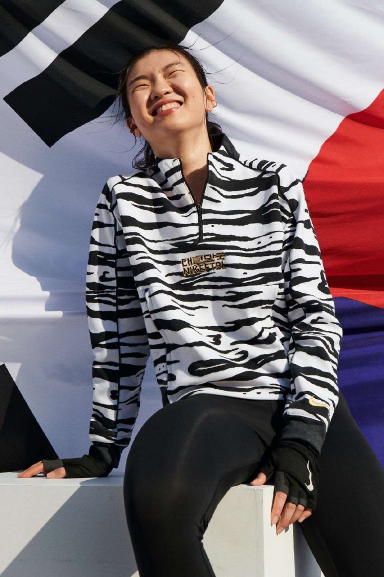 Smiling Asian woman poses in black-and-white tiger-stripe jacket, black leggings, and fingerless gloves against the Korean flag backdrop.