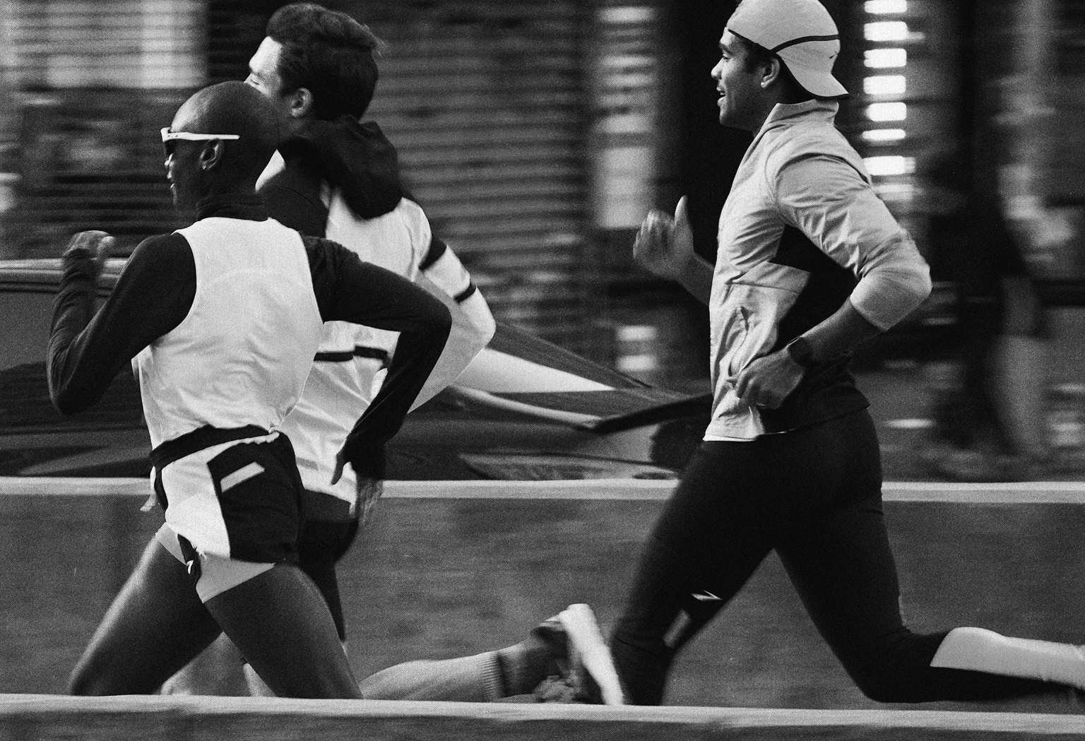 Three athletes run a race in a black and white photograph, showcasing speed and determination in a city setting with blurred background.