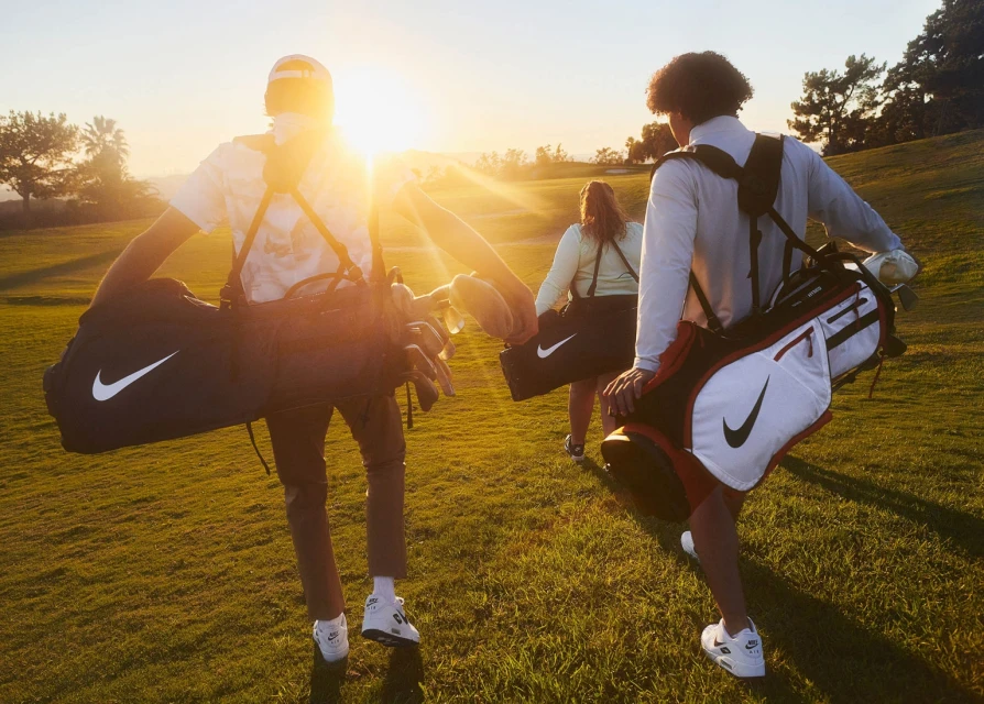 Three people walk across a lush green golf course at sunset, carrying Nike golf bags and sporting athletic wear and casual attire.