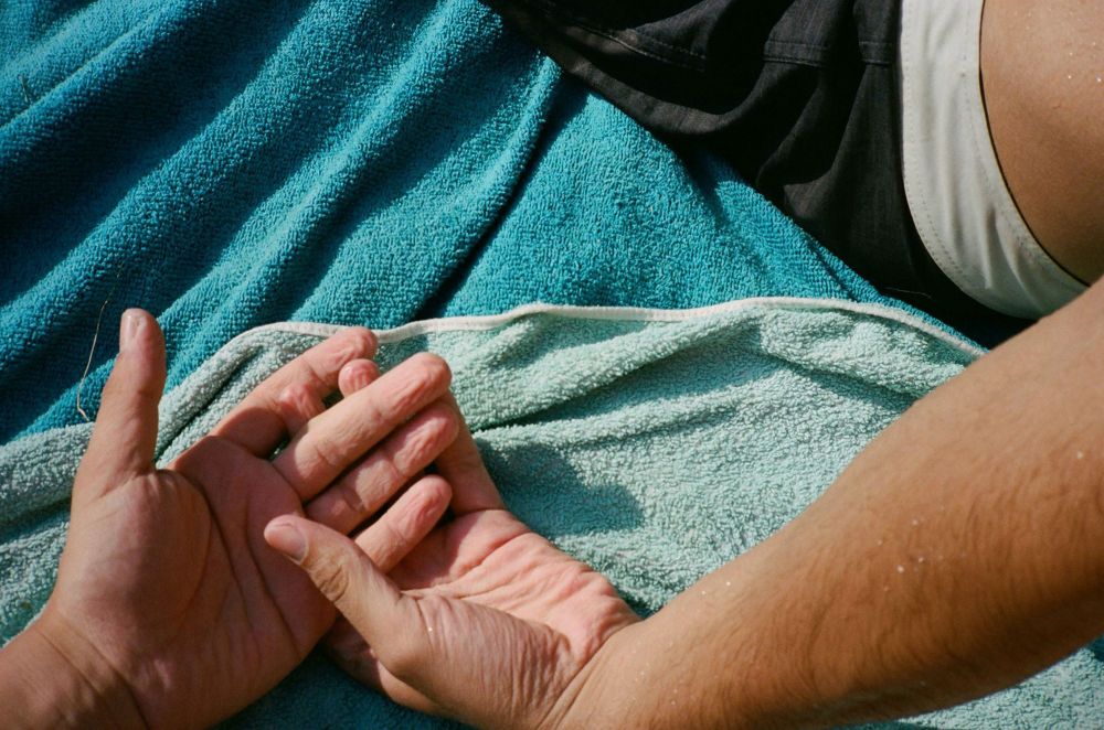 Close-up overhead shot of two light-skinned adult hands intertwined on a turquoise beach towel near black shorts.