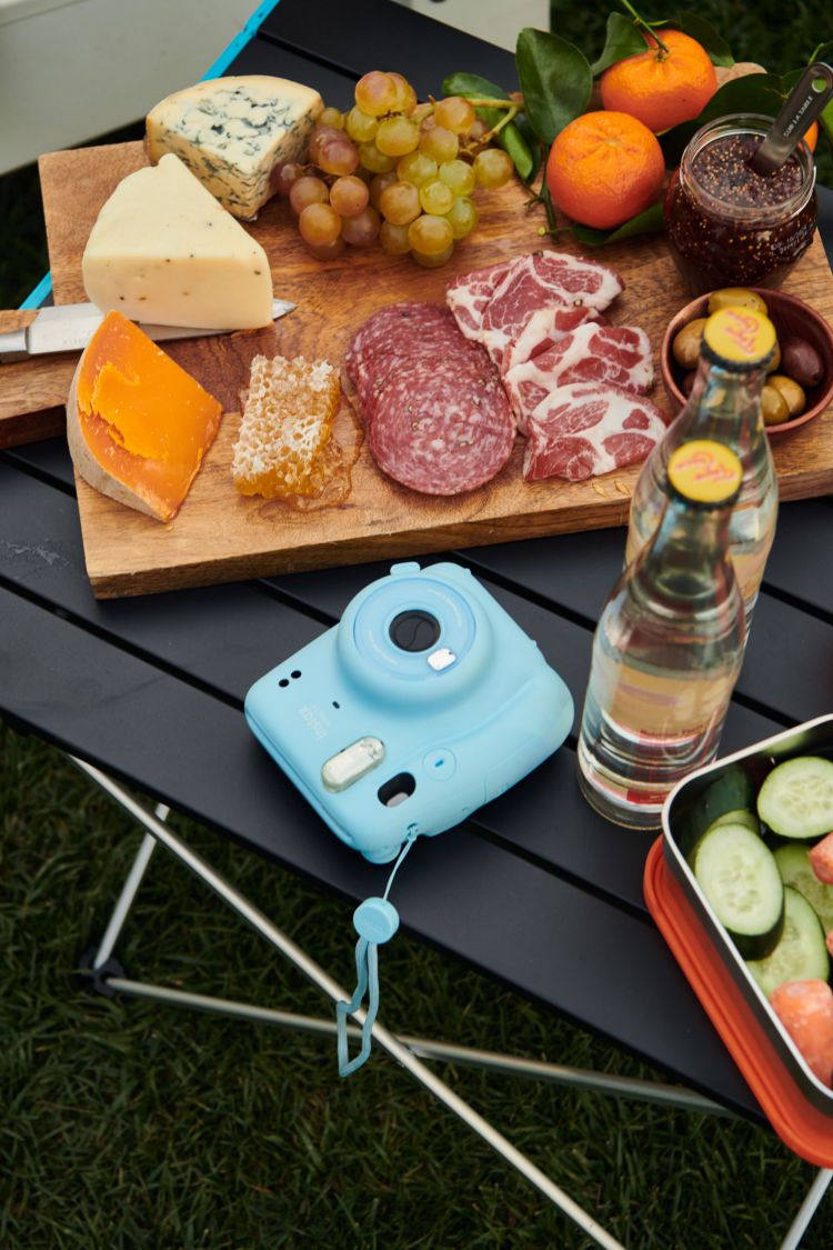 Overhead shot of an Insta camera, assorted cheeses, cured meats, and fruits on a wooden board atop a black folding table on grass.