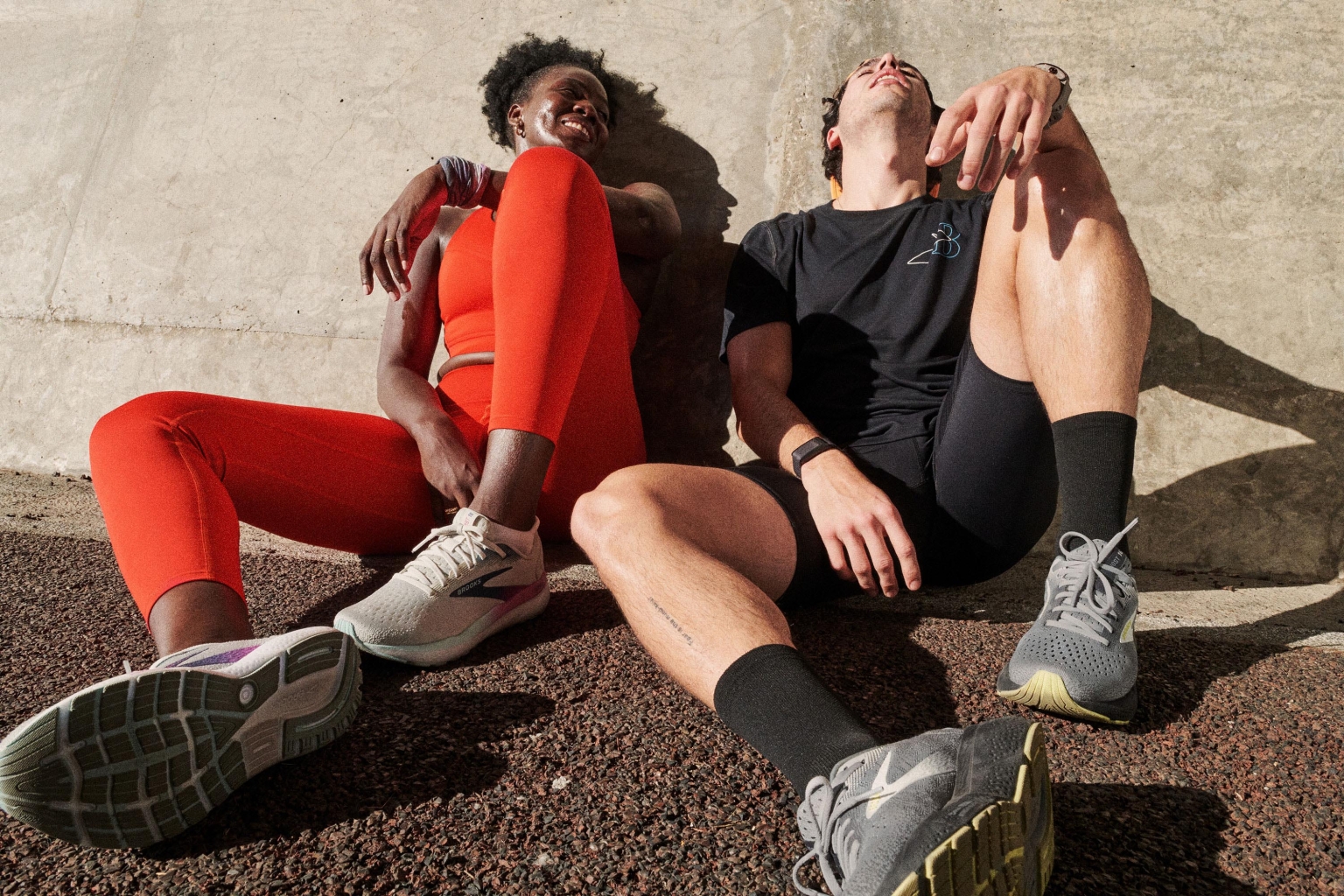 Two athletes in fitness apparel resting outdoors; the woman is smiling, wearing an orange top and leggings; the man is wearing black activewear.