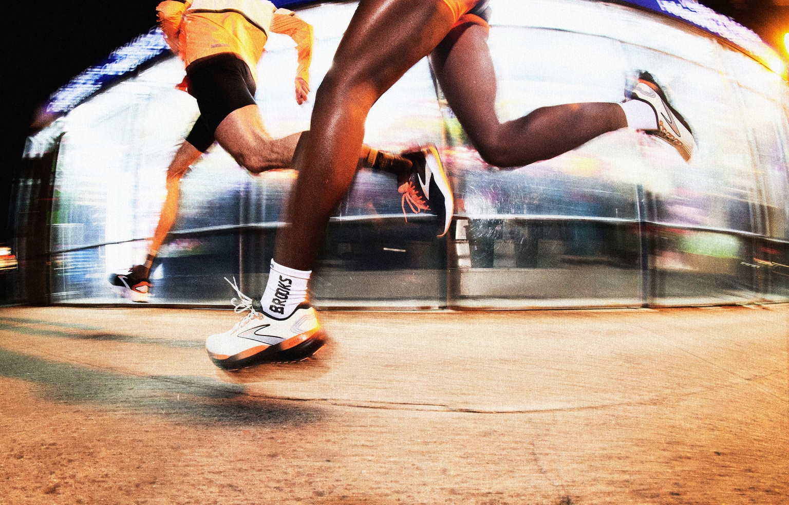 Two runners sprint on a city street at night, captured with motion blur. Wearing running shoes, shorts, and athletic gear under bright streetlights.