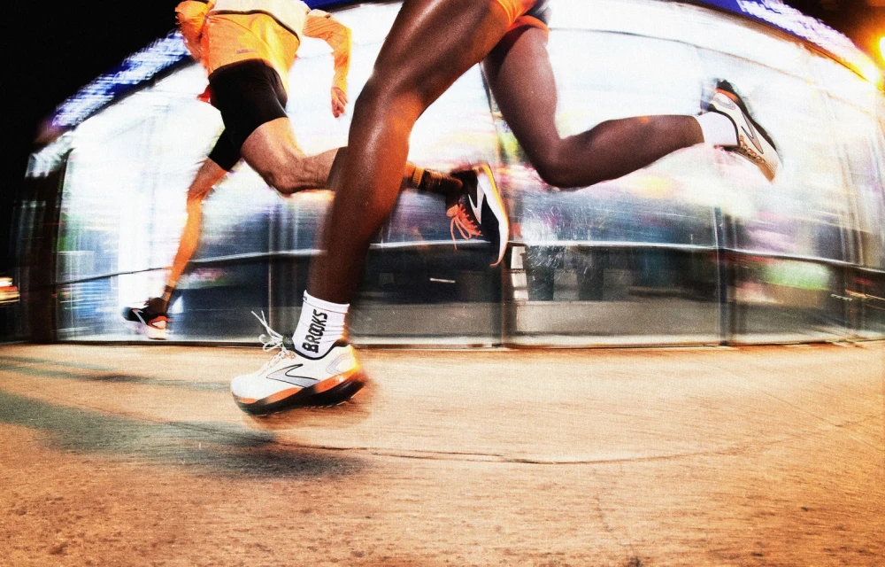 Two runners sprint on a city street at night, captured with motion blur. Wearing running shoes, shorts, and athletic gear under bright streetlights.