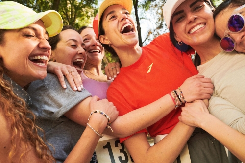 Group of smiling women in athletic wear embrace outdoors near a race marker. Women wear caps and athletic shirts, exhibiting joy in daylight.