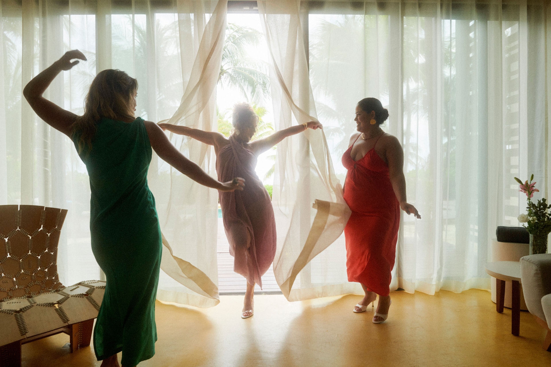 Three women in dresses pose and dance near a window with sheer curtains, with sunlight streaming through, creating a bright and airy atmosphere.