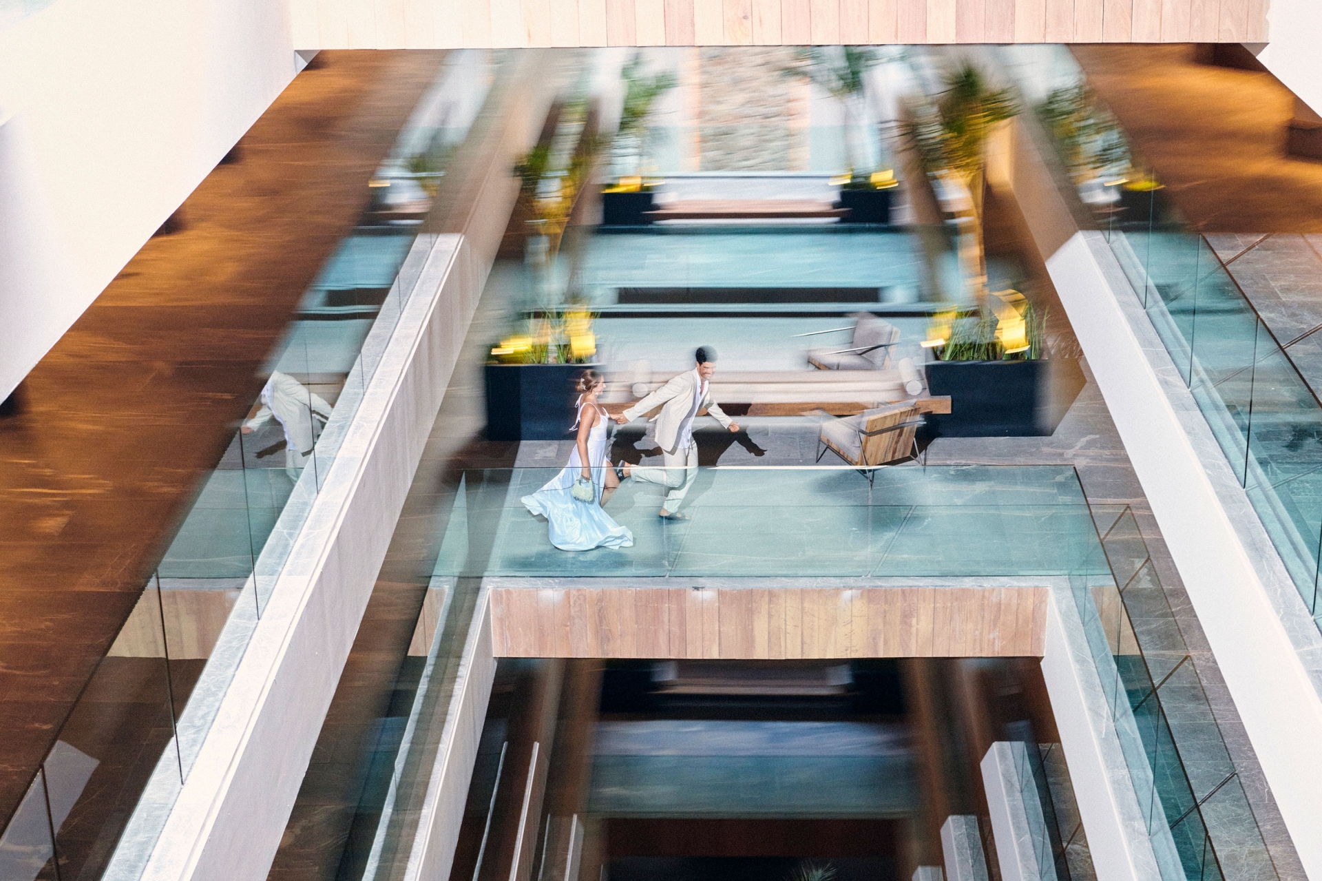 Aerial view of a couple in formal wear holding hands and running across a glass walkway in a modern, multi-level building with wooden floors.