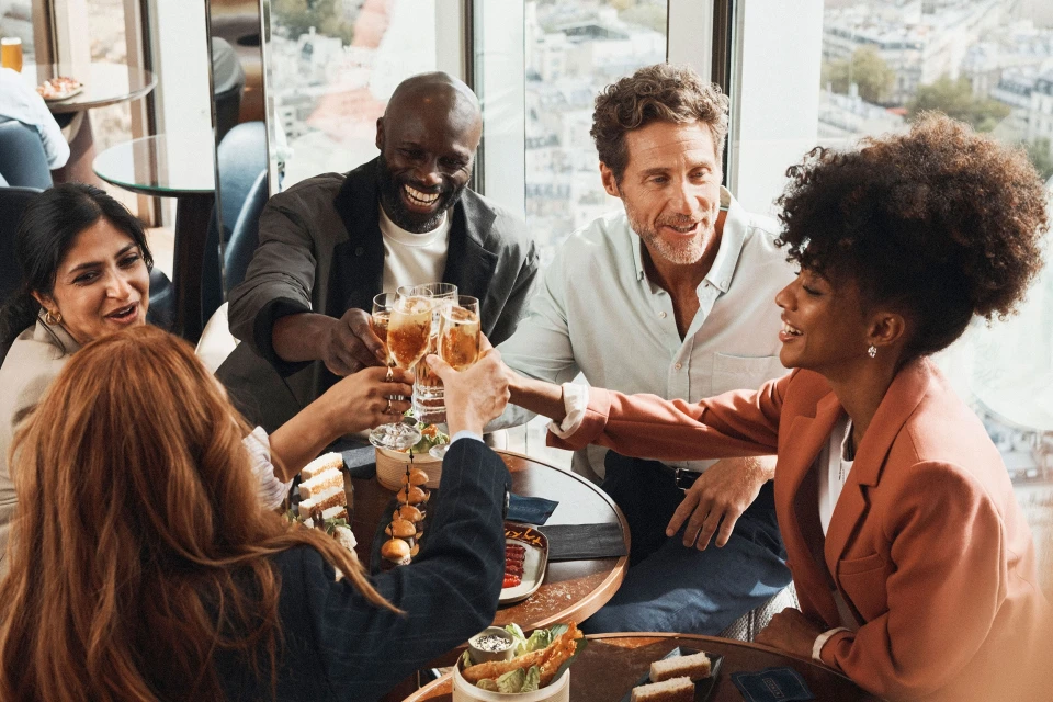 Five smiling adults raise champagne flutes in a celebratory toast around a low table in an indoor venue with a city view.