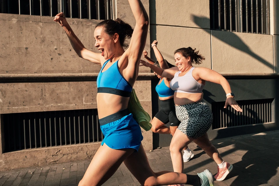 Three women in athletic wear are running outside, laughing with raised arms, and exercising in sunlight, showcasing fitness and active lifestyle.