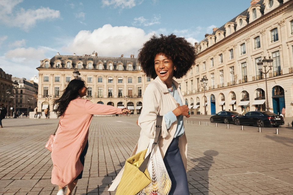 Two stylish women stroll through a city square, laughing and enjoying the day with historic buildings and blue sky in the background.