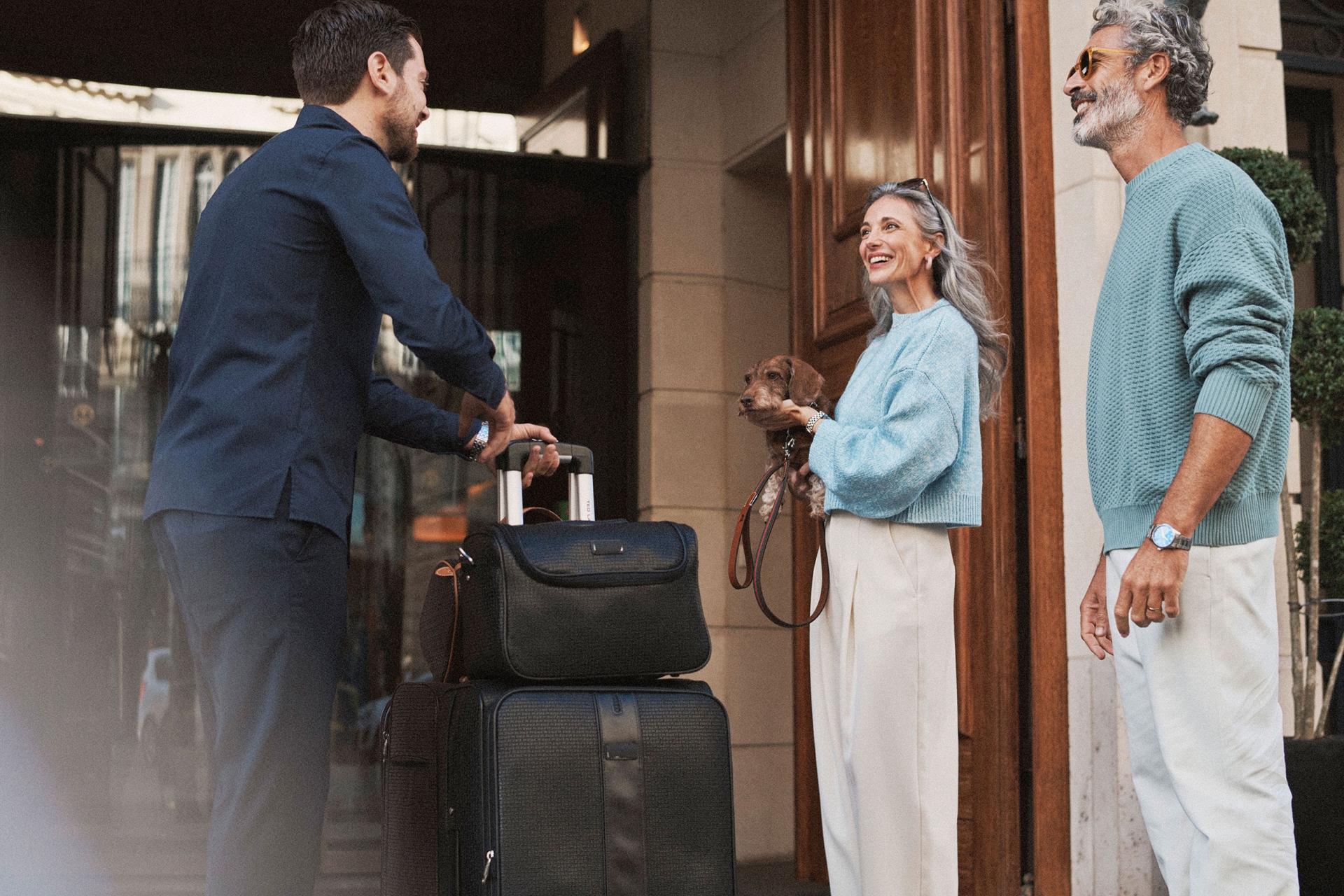 Hotel porter assisting a mature couple with luggage and a dog outside a building, demonstrating upscale travel and customer service.