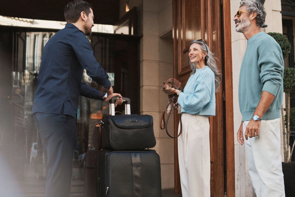 Hotel porter assisting a mature couple with luggage and a dog outside a building, demonstrating upscale travel and customer service.