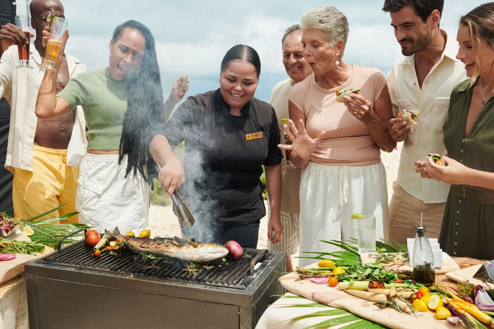 Outdoor BBQ scene featuring a chef grilling fish for a group of diverse adults on a sunny day, showcasing food preparation and social gathering.