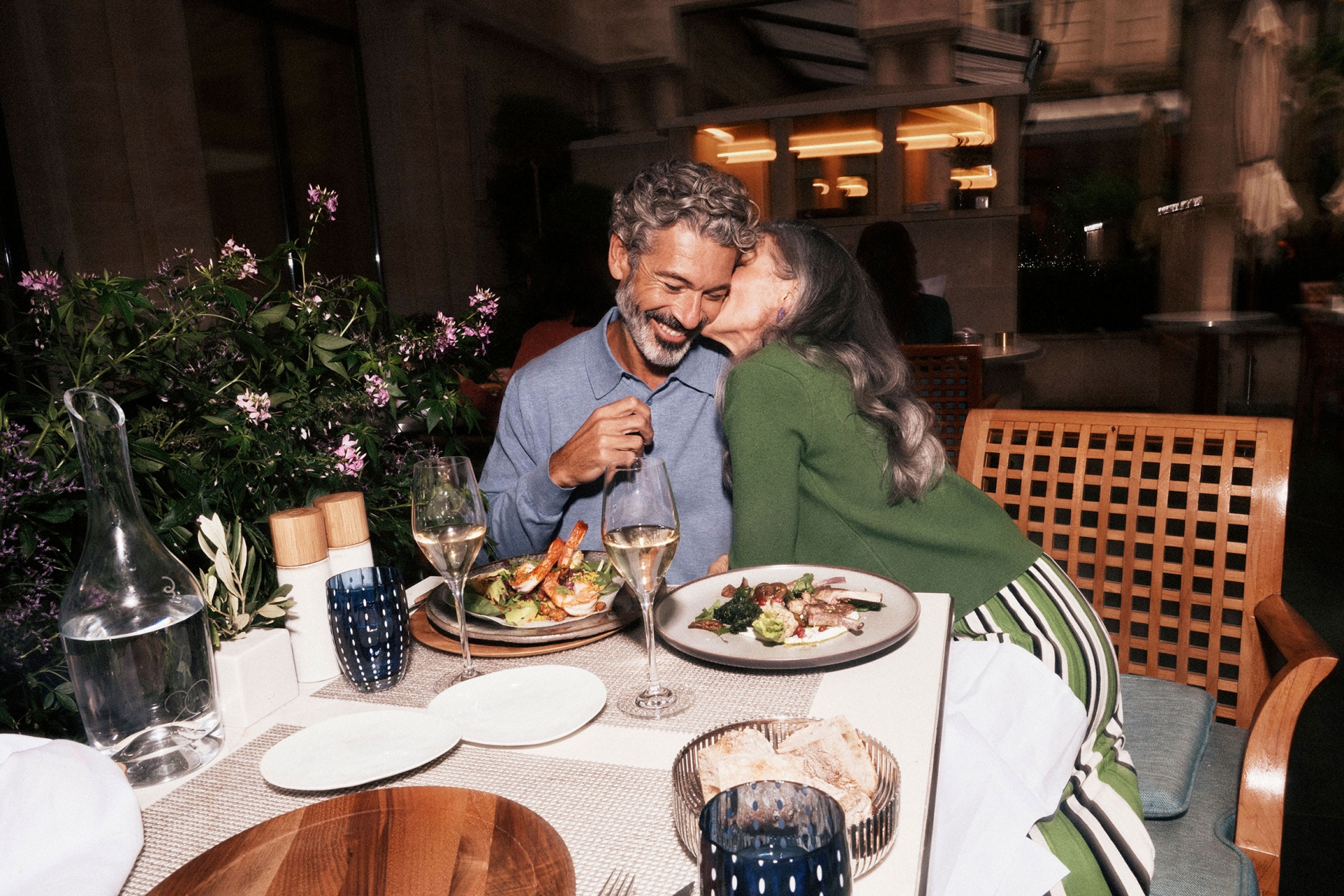 A grey-haired couple dines at an upscale restaurant, sharing a kiss and enjoying wine with appetizers. The atmosphere is intimate and well-lit.