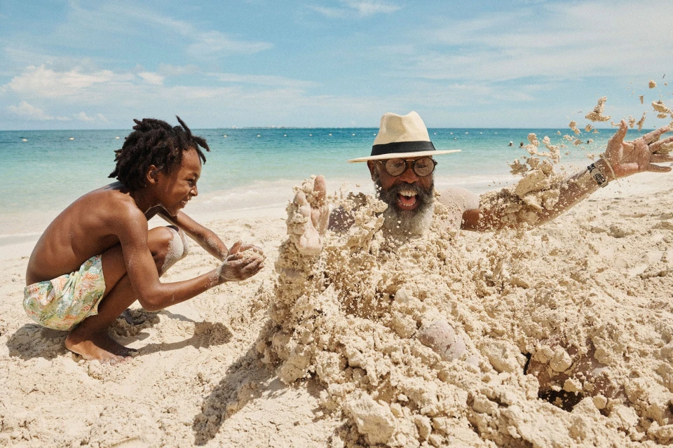 Child playing in the sand with an adult buried up to the chest on a sunny beach with clear blue ocean and sky in the background.