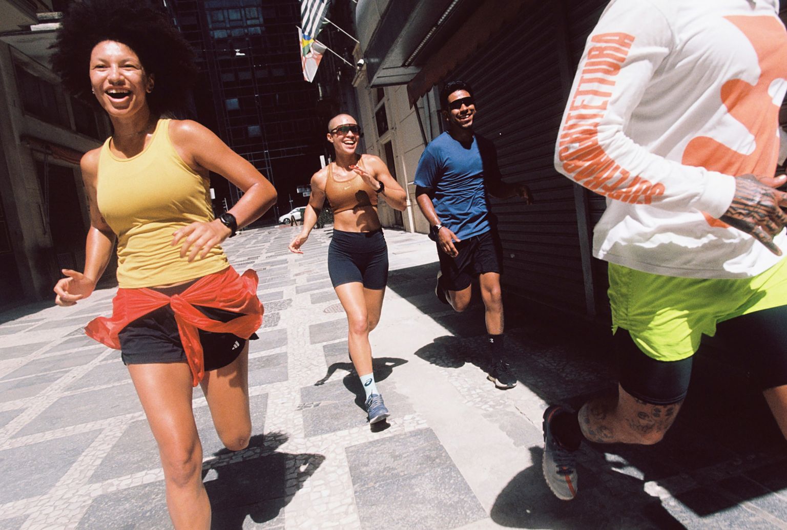 Four athletic adults running on a city street during daylight, wearing activewear and fitness trackers, smiling and looking ahead.