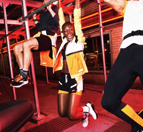 Three fit adults doing pull-ups on metal scaffolding. A woman in the foreground is smiling, wearing fitness apparel and yellow jacket.