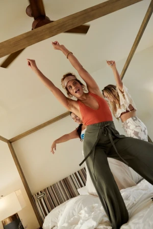Three adult women on a bed raise their arms in celebration, view from below; warm-toned bedroom with a ceiling fan and white linens.