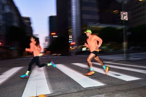 Two athletic adults in running gear sprint across a crosswalk in a city at dusk, buildings blurred in the background.