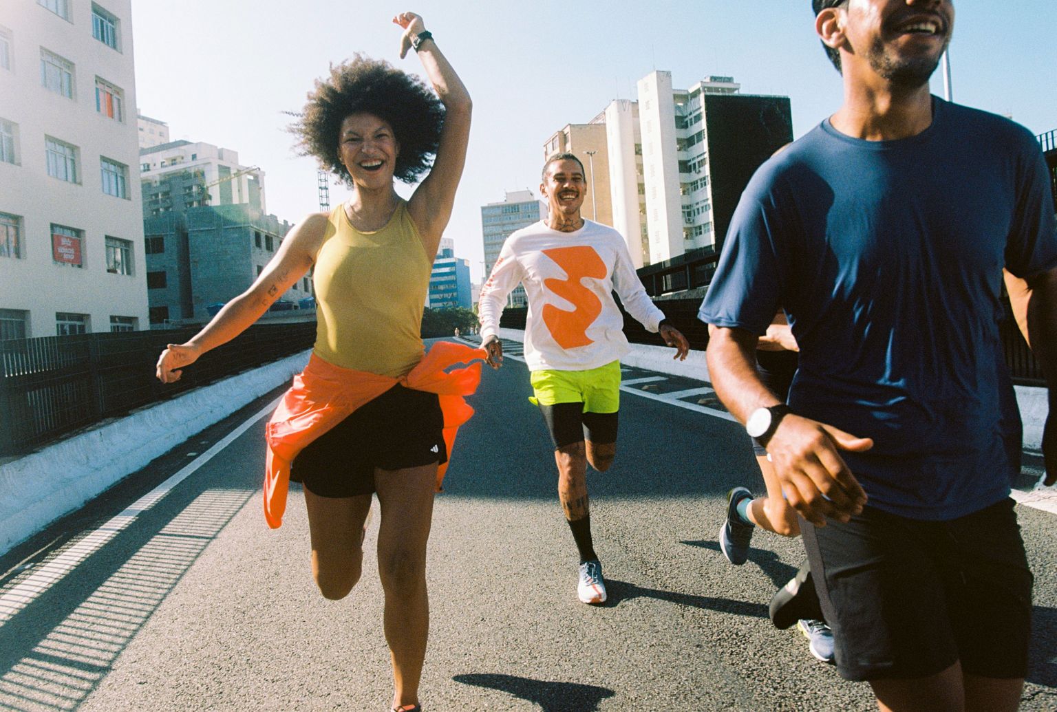 Three adult athletes run toward the camera on an asphalt road in an urban environment on a sunny day. Fitness and sport.