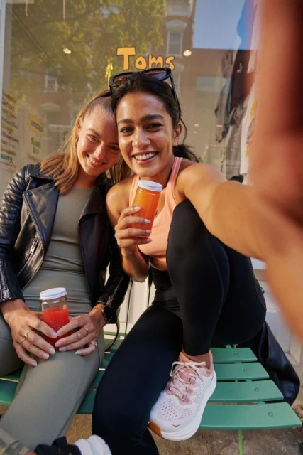 Two smiling women pose with juice outdoors. One wears a leather jacket; the other has sunglasses on her head, sitting on a green bench.
