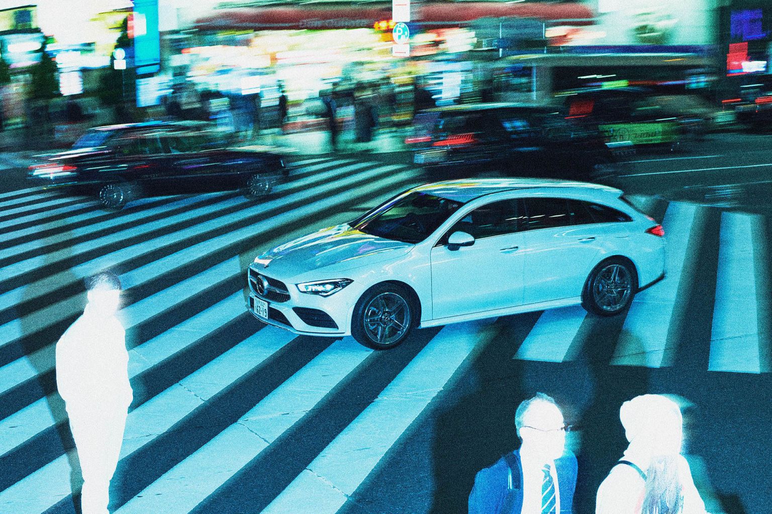 White Mercedes-Benz wagon on a crosswalk at night with blurred pedestrians and other vehicles in a motion-blurred urban setting.