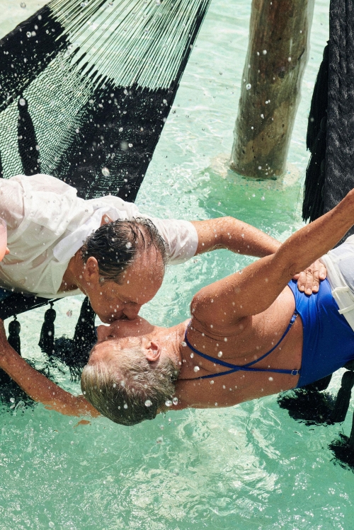 A couple embraces and kisses in a black hammock suspended above clear, shallow water with a wooden support structure nearby.