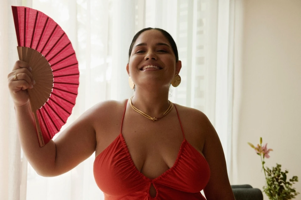 Smiling woman in a red dress poses indoors, holding a red hand fan. She wears gold earrings and a necklace, looking toward the camera.