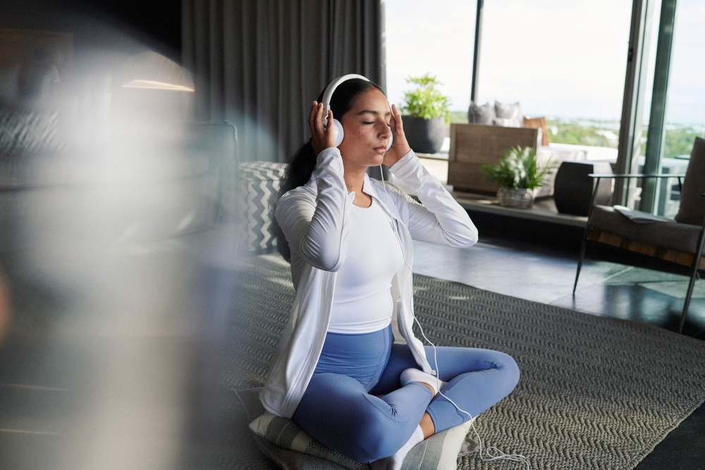 Young woman wearing headphones sits cross-legged indoors, eyes closed, white athletic jacket, blue leggings, relaxing in tranquil home setting.