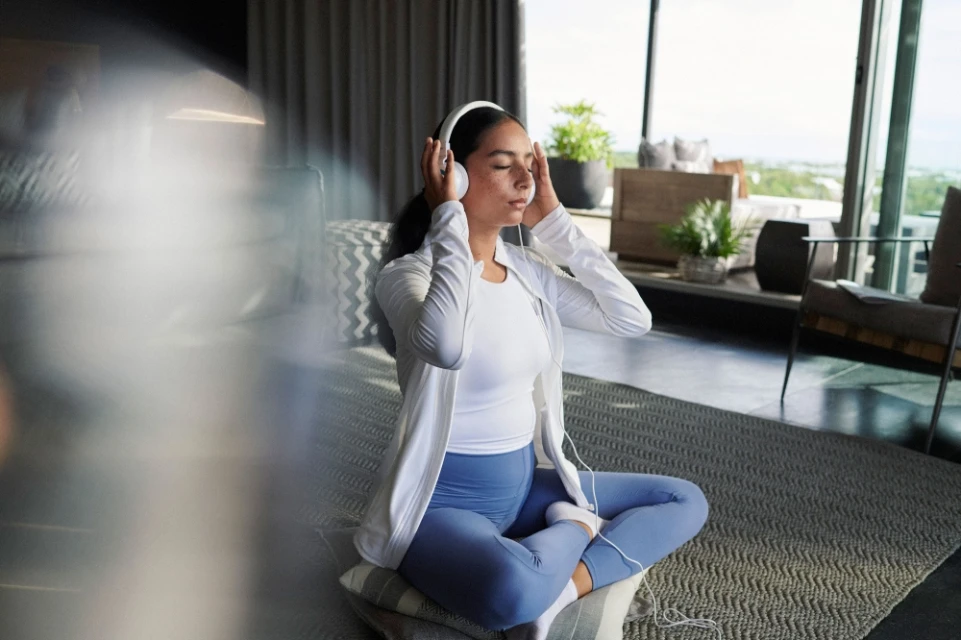Young woman wearing headphones sits cross-legged indoors, eyes closed, white athletic jacket, blue leggings, relaxing in tranquil home setting.