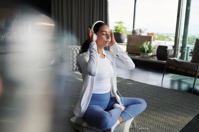 Young woman wearing headphones sits cross-legged indoors, eyes closed, white athletic jacket, blue leggings, relaxing in tranquil home setting.