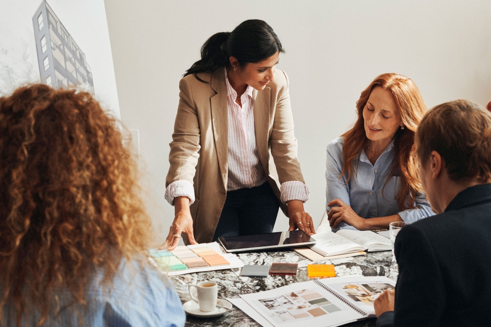 A collaborative business meeting with four people gathered around a table, discussing architectural plans and reviewing color samples.