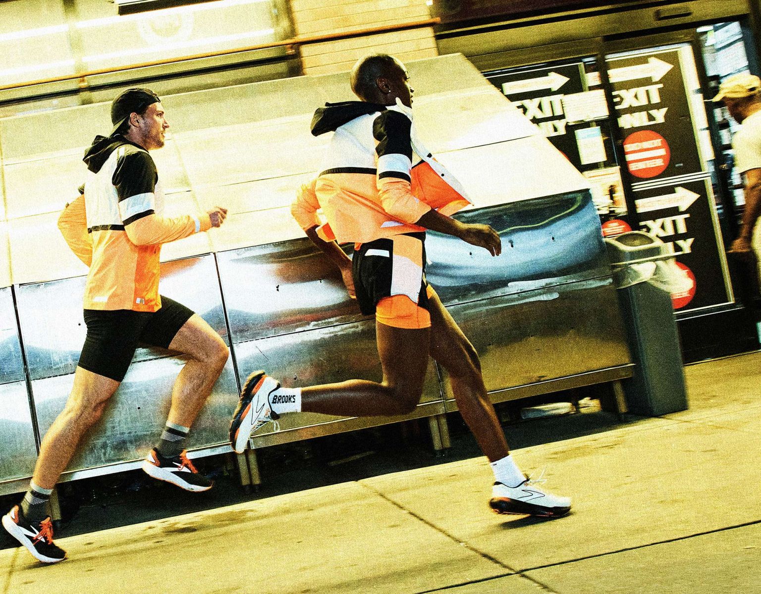 Two runners, an adult male and female, wearing athletic apparel, run outdoors on a city street, with a metal structure behind them.
