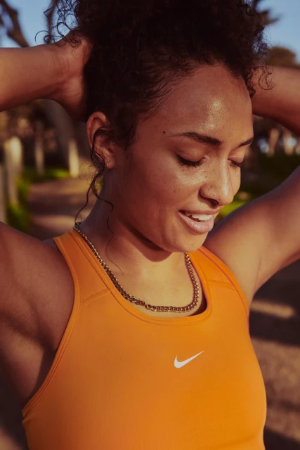 Athletic woman in orange sports bra pauses during outdoor workout to adjust hair, showcasing fitness and healthy lifestyle.