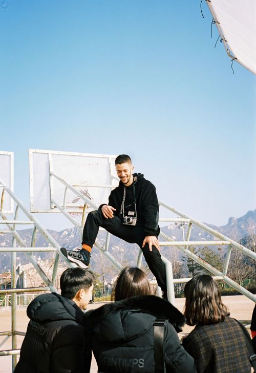 A man stands on a basketball hoop as three people look up at him in a candid outdoor shot on a clear day with mountains in the background.