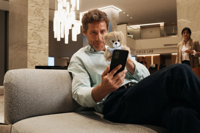 Man looks at a smartphone while sitting on a couch with a teddy bear in a bright hotel lobby near a woman walking in the background.