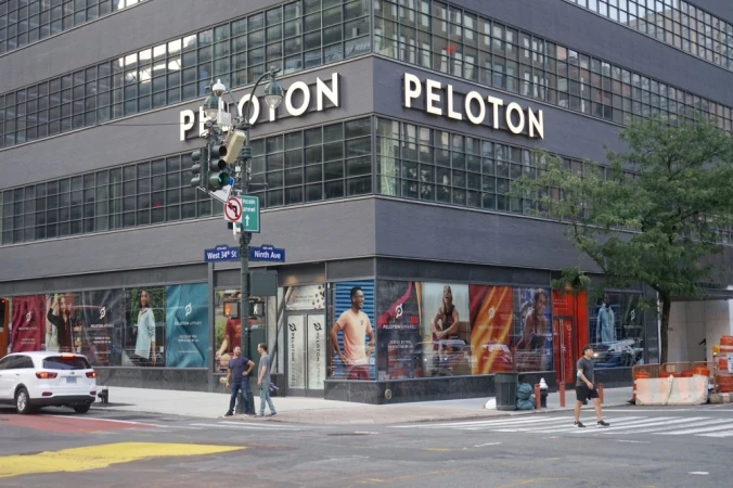 Peloton store exterior in New York City. Three pedestrians cross the street in front of the storefront windows and a white car drives by.