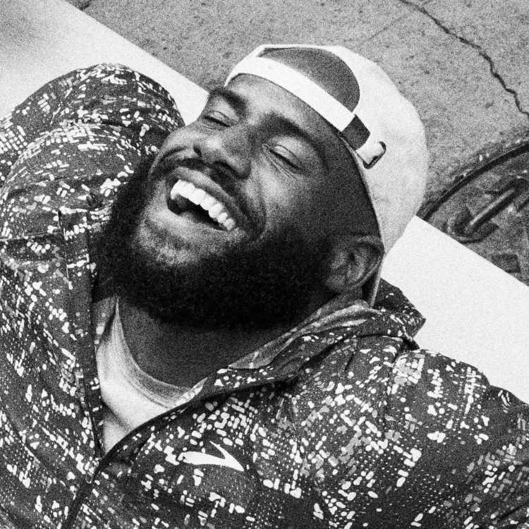 Black man with a beard wearing a baseball cap and patterned athletic jacket laughing while lying down