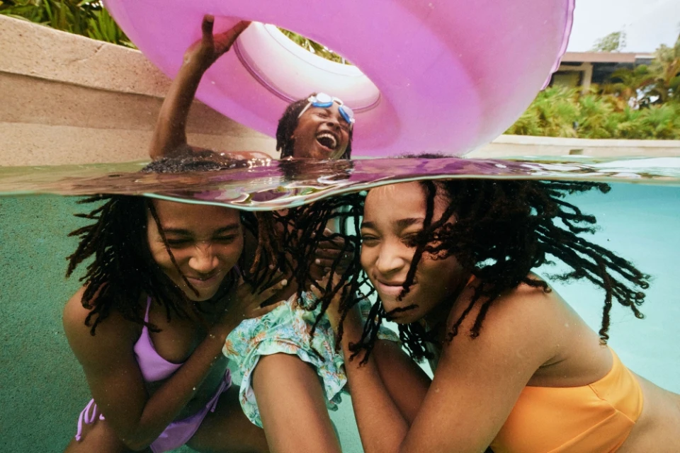 Three African American women laughing in an outdoor swimming pool; one holds a pink inflatable ring, captured with a medium shot.