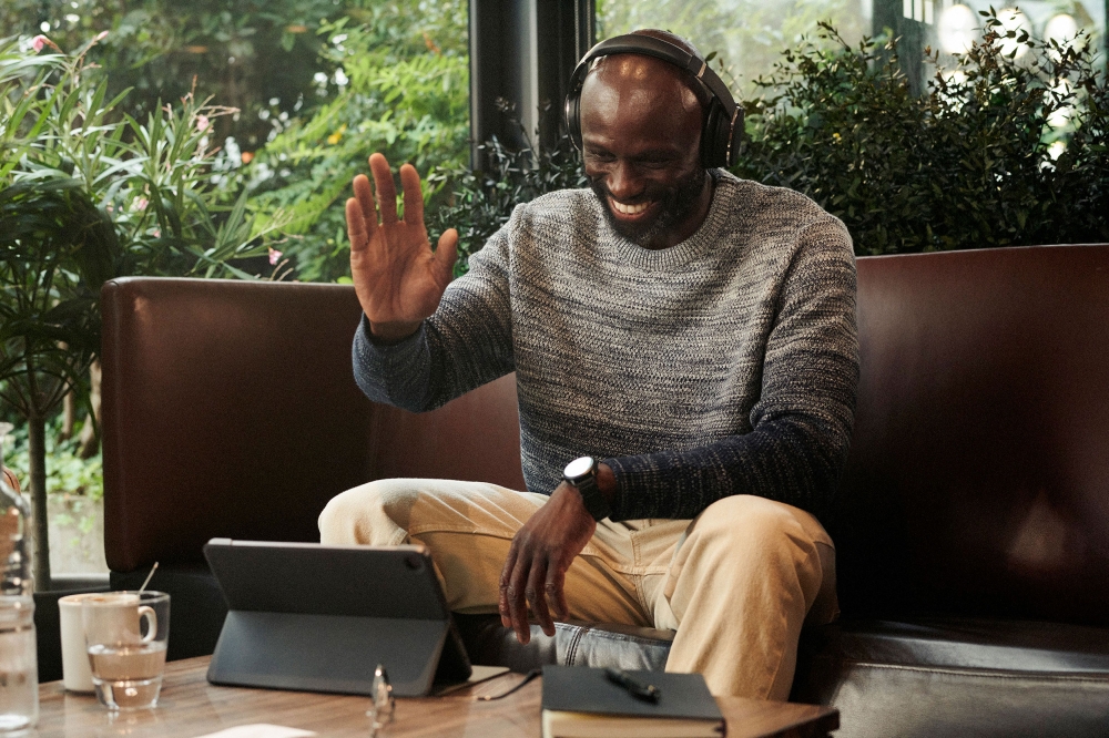 Adult male with headphones on video call using a tablet, waving while sitting on a brown couch in a bright indoor setting.