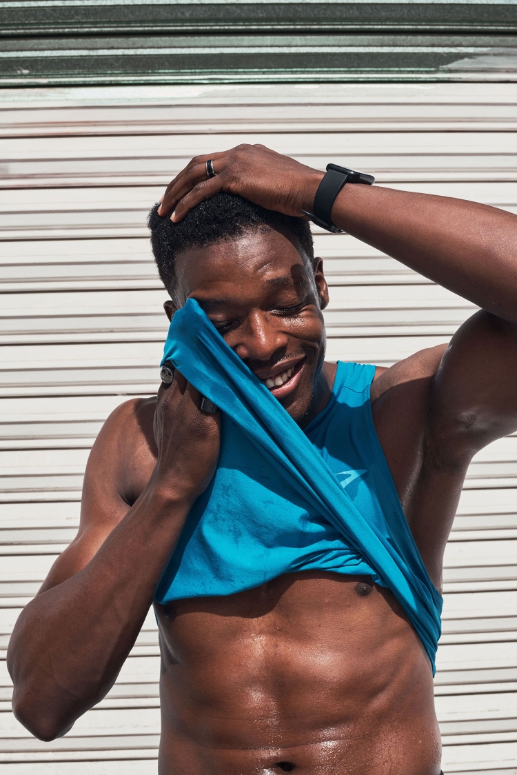 Athletic Black man smiles while wiping sweat with a blue shirt in front of corrugated metal. Workout, fitness, exercise.