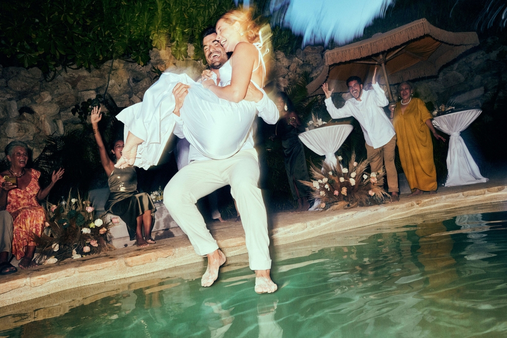 Man carries woman toward pool, wedding guests celebrate in background. Outdoor party, casual attire, candid moment, shallow depth of field.