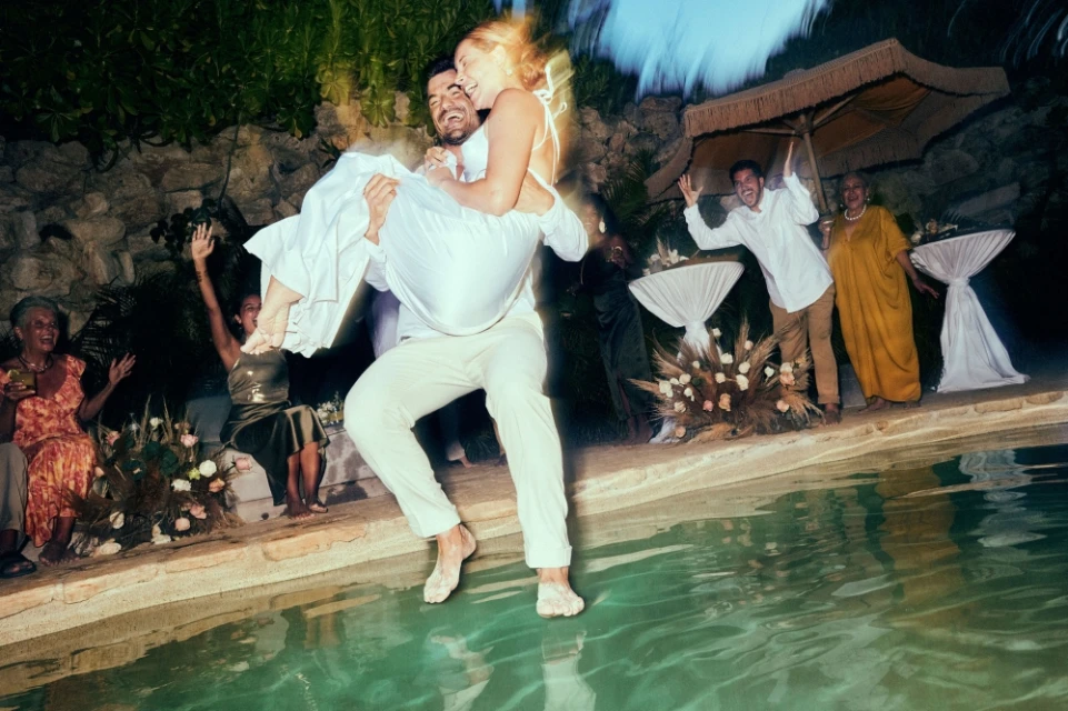 Man carries woman toward pool, wedding guests celebrate in background. Outdoor party, casual attire, candid moment, shallow depth of field.