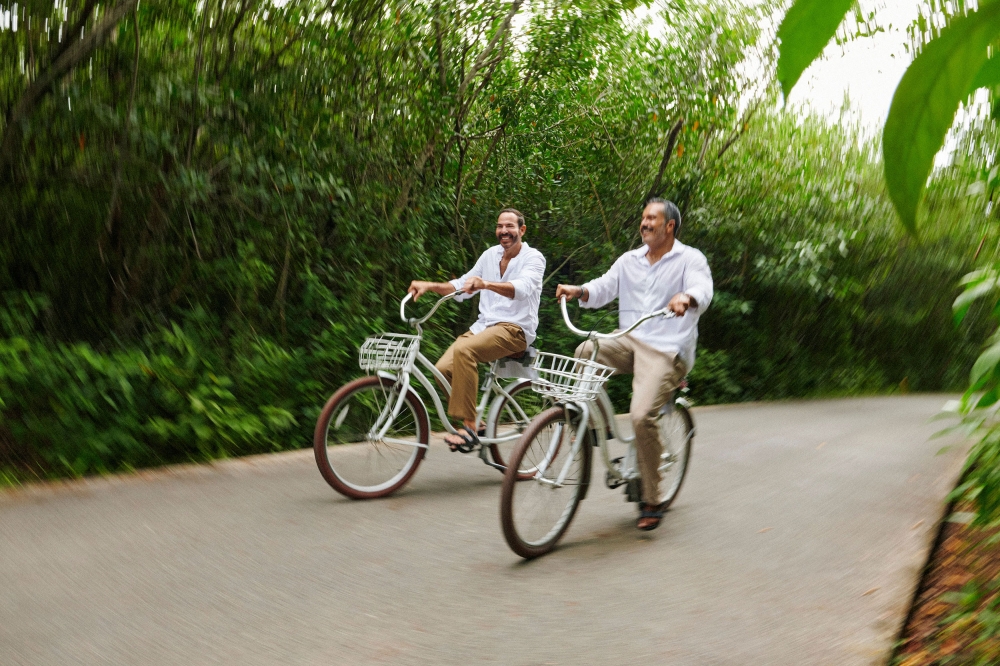 Two men in casual summer clothing ride beach cruisers along a winding road lined with lush green trees and foliage in an outdoor setting.