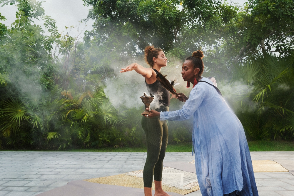 Woman receives smoke cleansing from another woman in outdoor setting with lush trees, creating a serene and holistic wellness scene.