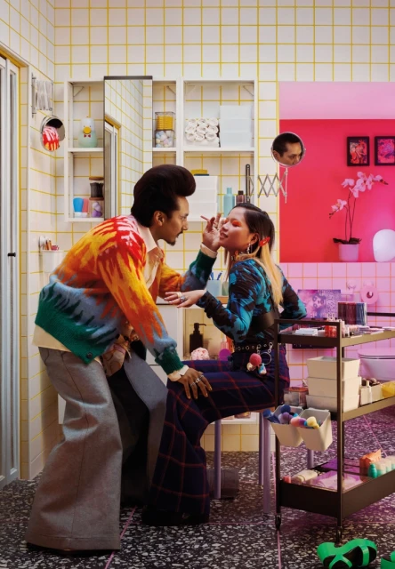 Two people in a yellow-tiled bathroom. One person applies makeup to another seated on a metal stool next to a metal shelving unit.