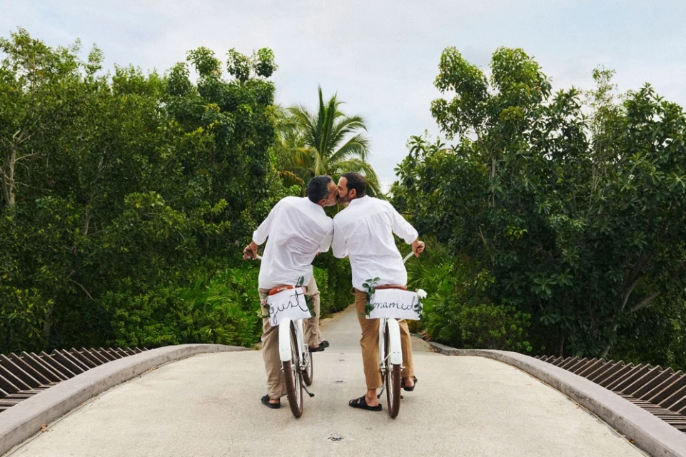 Two adult males on bicycles kiss on a bridge surrounded by green foliage, wearing white shirts and tan pants with 'Just Married' signs.