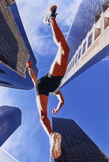 Low-angle shot of an athletic woman leaping in the air between tall buildings, wearing activewear and athletic shoes against a blue sky.
