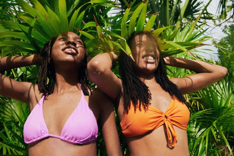 Two women in swimsuits smile in bright sunlight. Lush green foliage fills the background, creating a vibrant, tropical atmosphere.