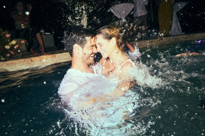 A happy couple in a pool embrace affectionately while water splashes around them at night during an outdoor celebration with guests.